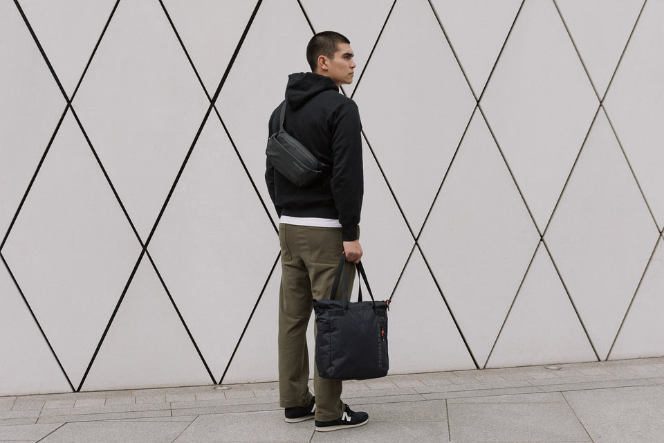 A man holding an All Black Ultra Light tote bag and wearing a black crossbody bag on his back