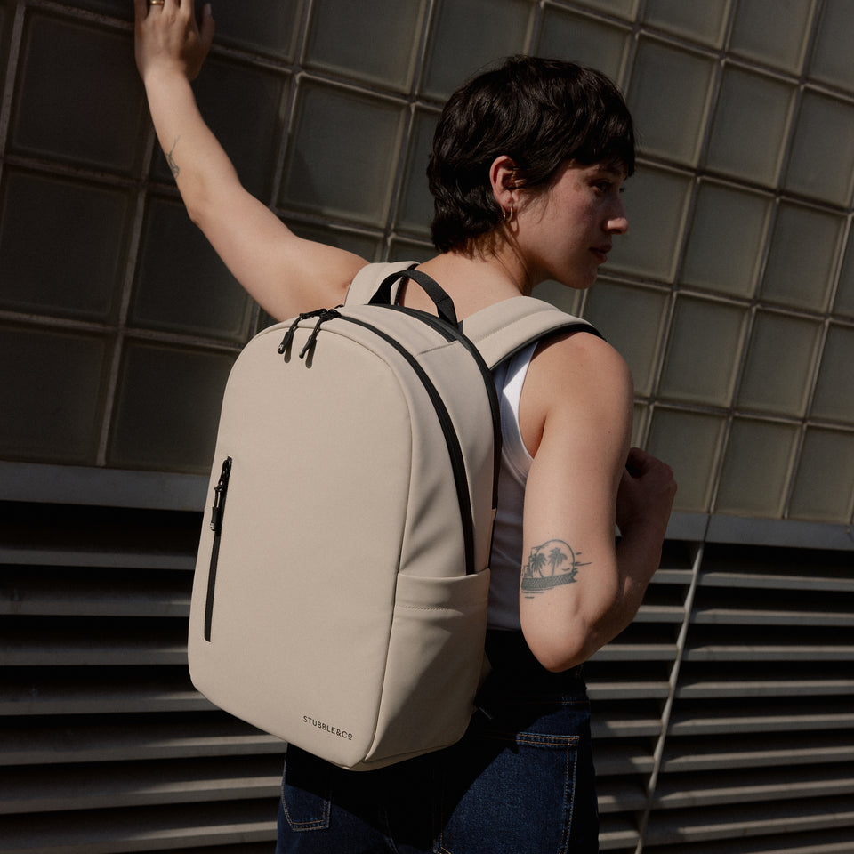 A woman wearing an Everyday Backpack 15L on her back in Sand as she leans against a wall